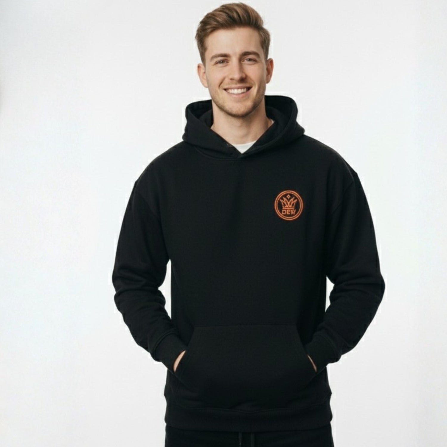 A male model smiling in a natural pose, wearing a premium black DEW hoodie with a circular orange brand logo on the chest, set against a clean white background.