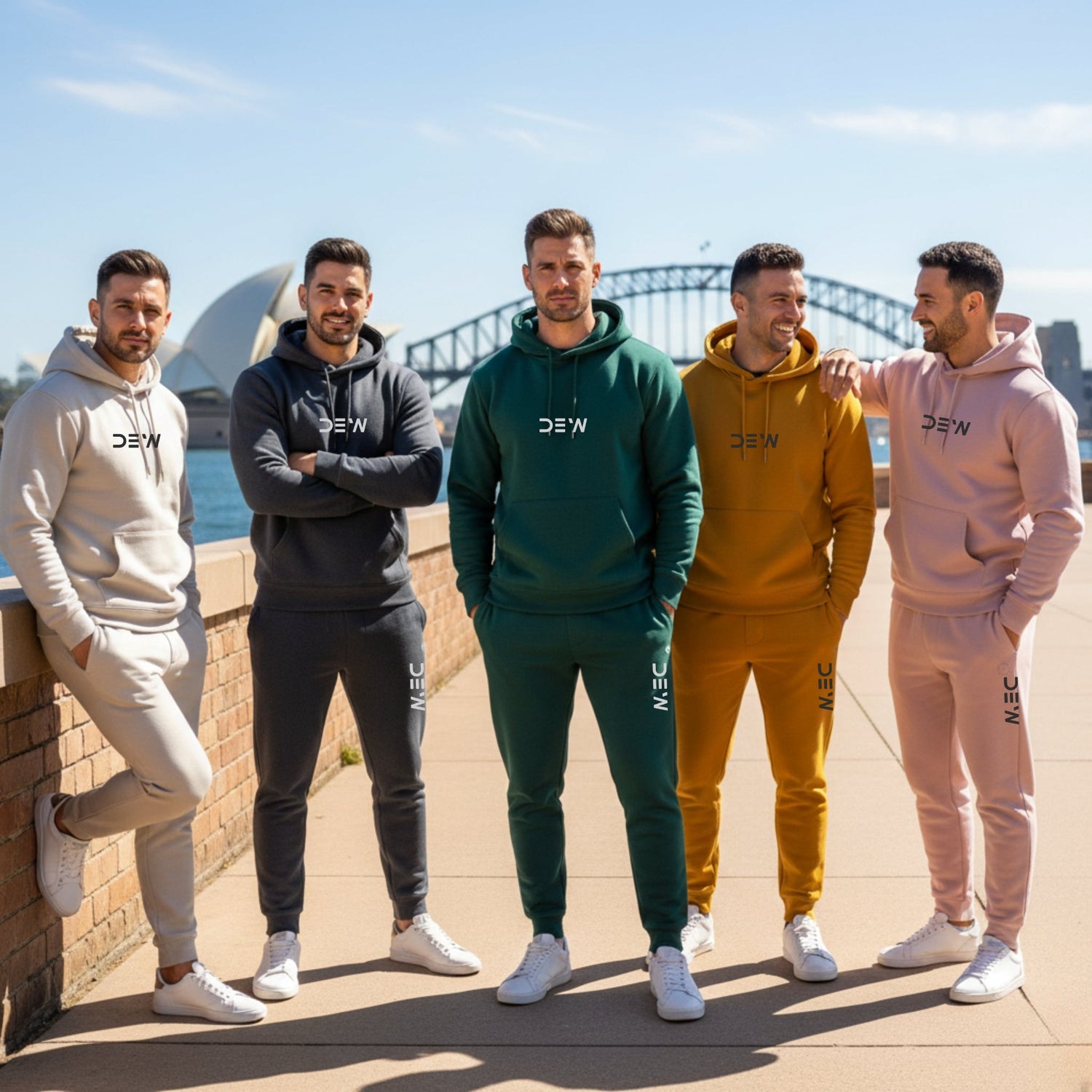 Five male models standing outdoors in front of the Sydney Harbour Bridge and Opera House, each wearing a matching, high-quality, unbranded hoodie and jogger set in shades of light grey, charcoal, dark green, mustard yellow, and pale pink.