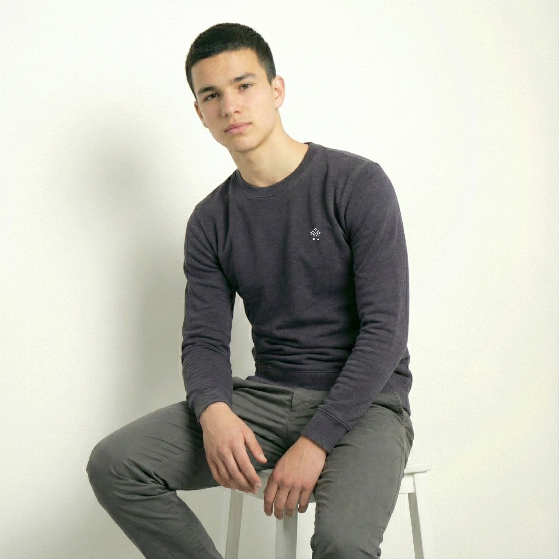 Person wearing a dark gray long-sleeve shirt and gray pants sitting on a white stool against a plain background.