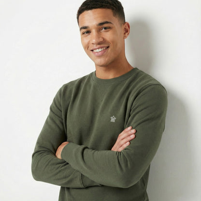 Man wearing a green sweatshirt with a small logo, standing against a white wall.