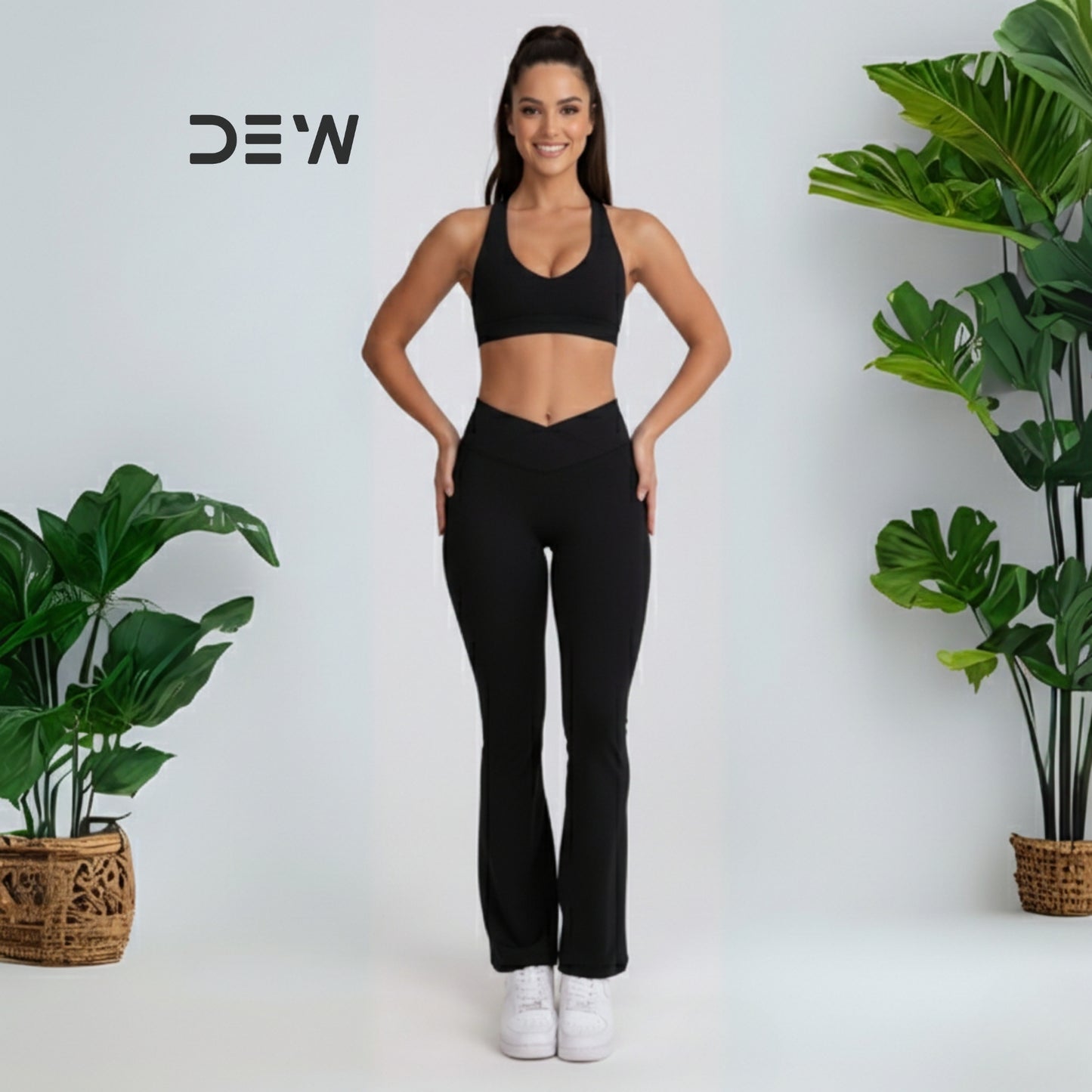 Woman in black athletic wear-flow fit flares standing between two potted plants with 'DEW' branding. 