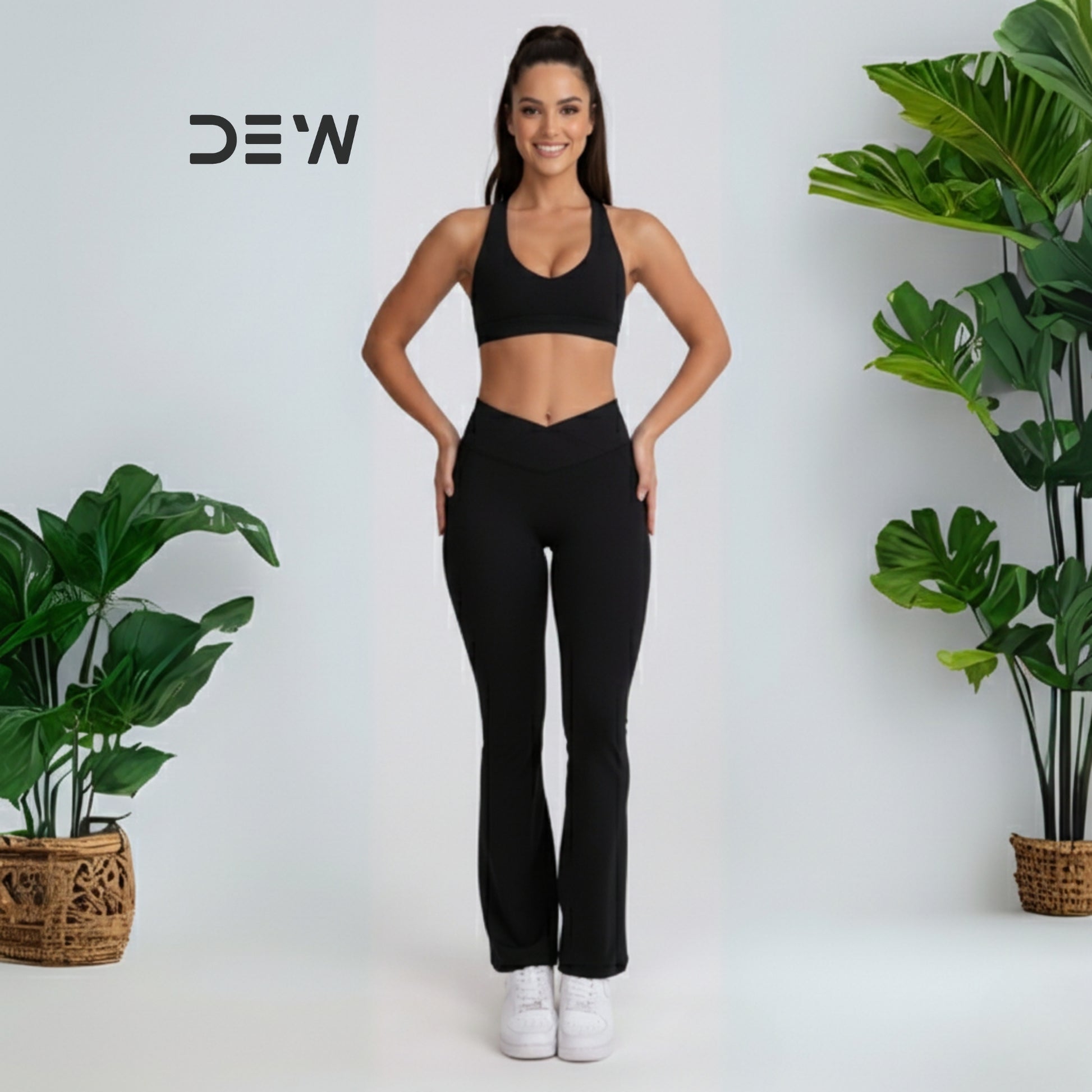 Woman in black athletic wear-flow fit flares standing between two potted plants with 'DEW' branding. 