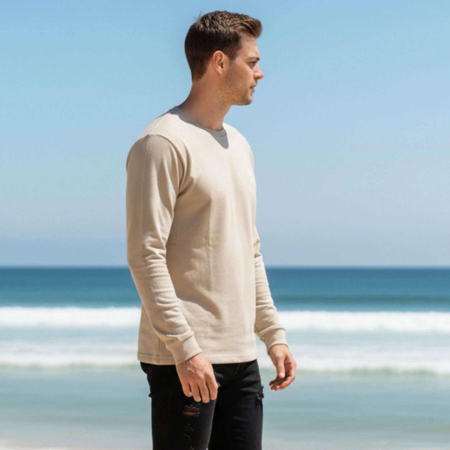 Man wearing a Dew 100% cotton beige t shirt standing on a beach with clear blue sky and ocean.