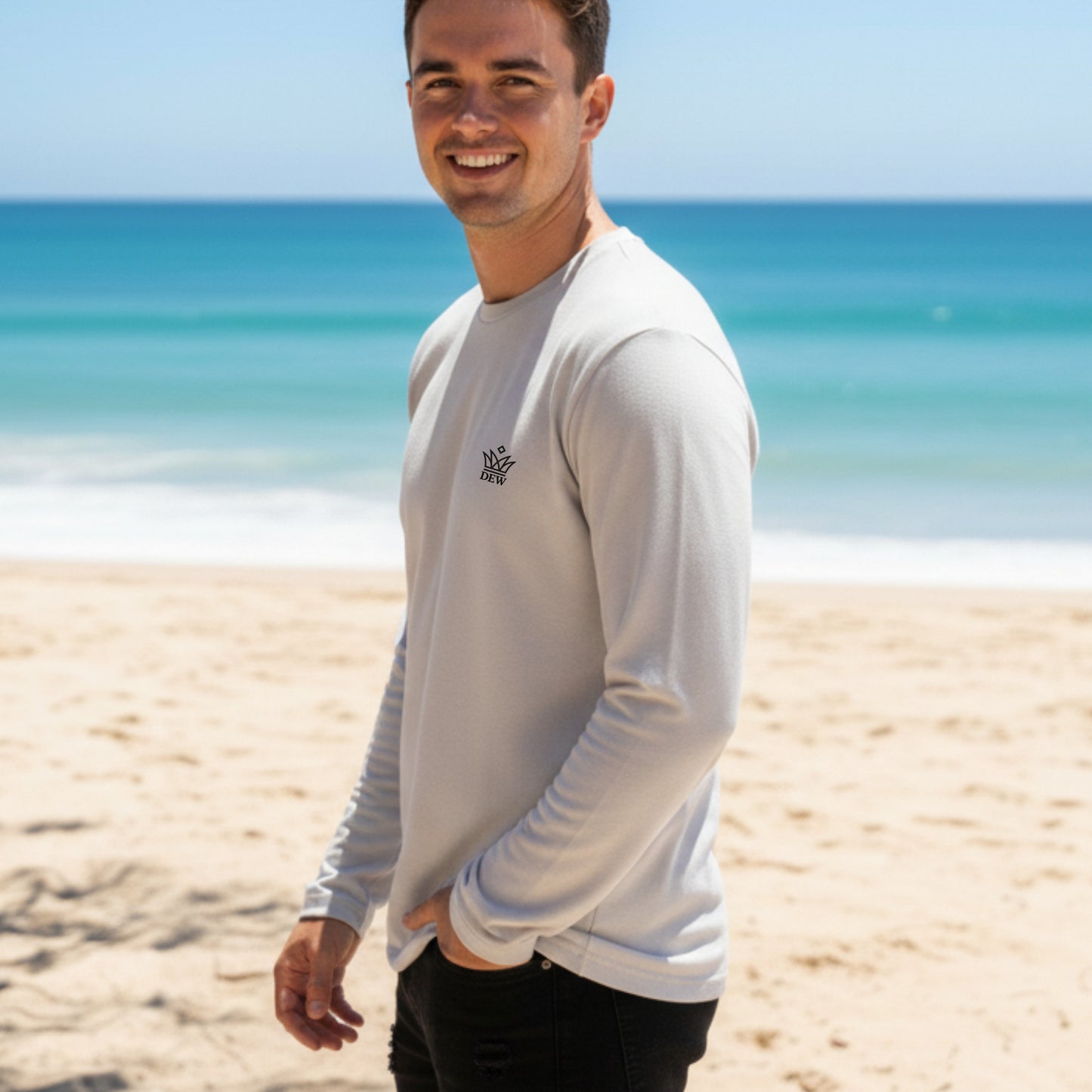 Man standing on a beach wearing a Dew 100 % cotton sea gray tshirt with a logo, smiling.