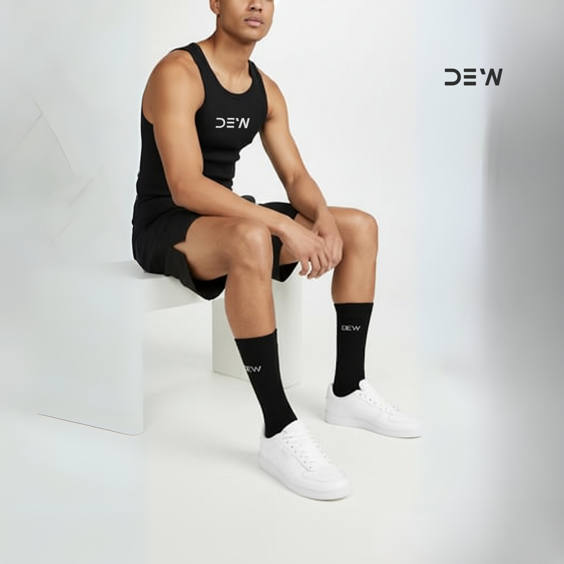 Person wearing black tank top, shorts, and socks with 'DEW' branding sitting on a white stool.