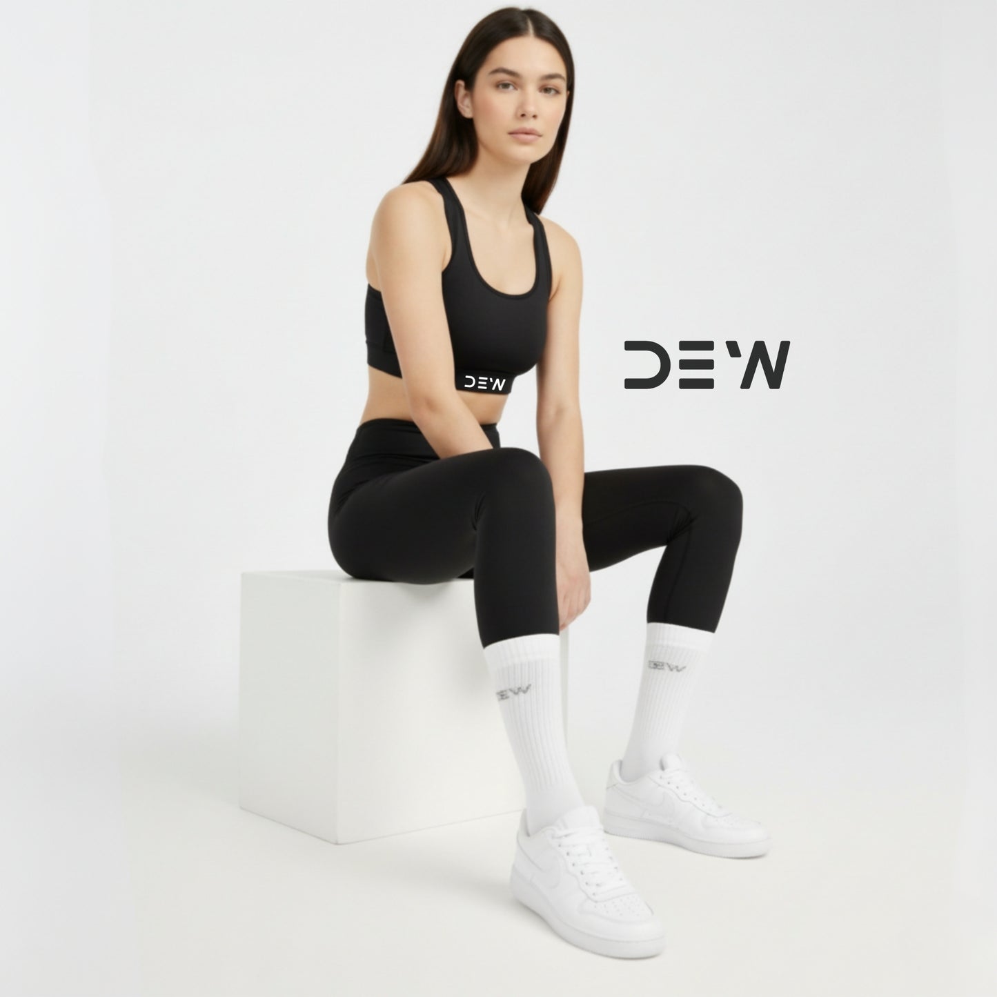 Woman in black sports outfit and bamboo socks with 'DEW' branding sitting on a white block against a light gray background.