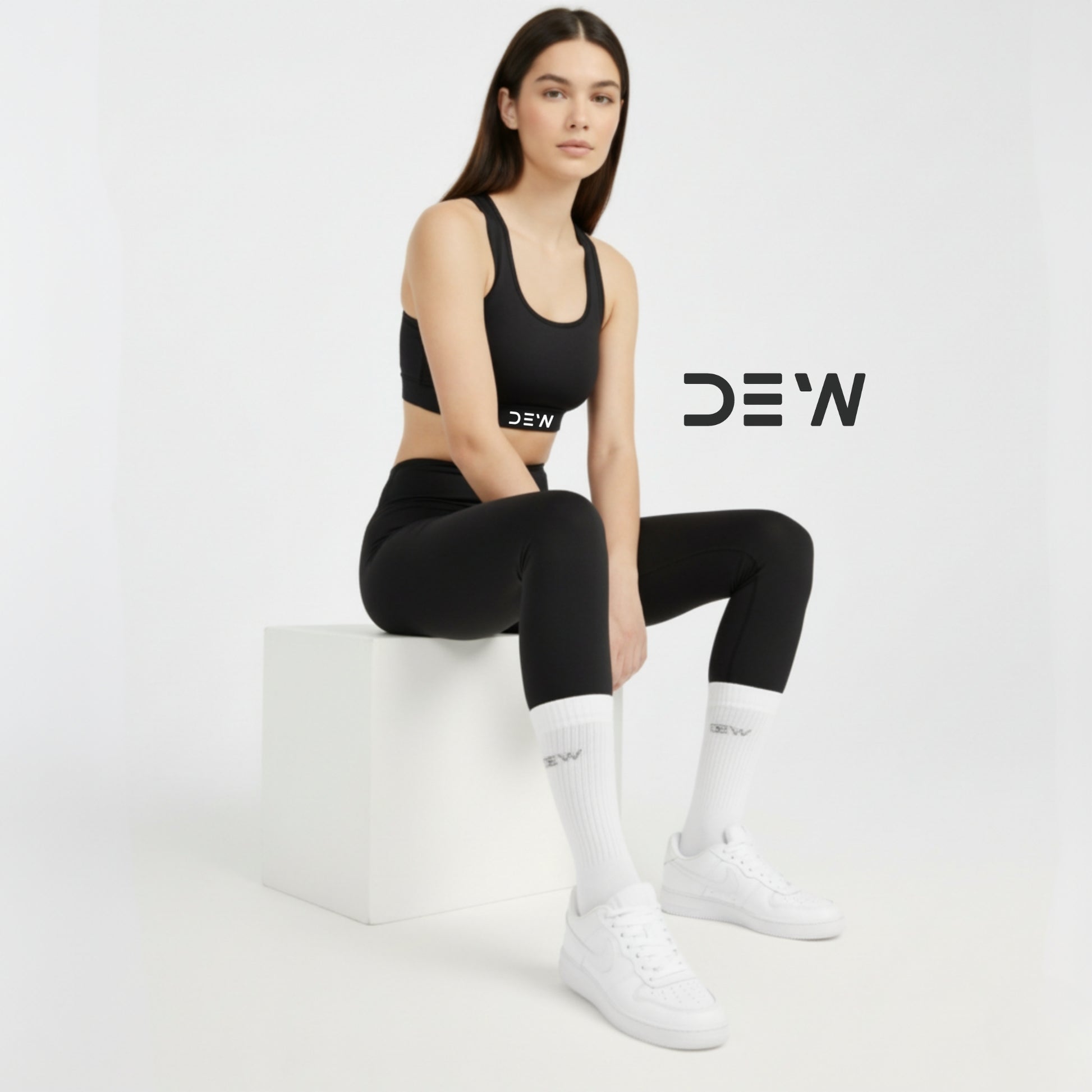 Woman in black sports outfit and bamboo socks with 'DEW' branding sitting on a white block against a light gray background.
