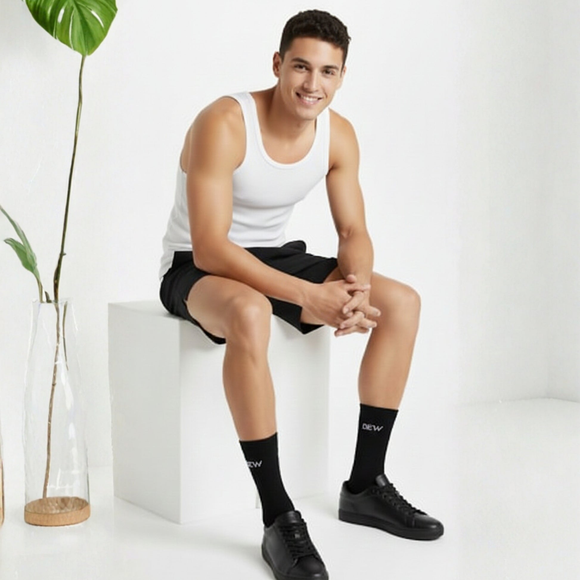 Man sitting on a white cube wearing a white tank top, black shorts, and black Dew socks with text.
