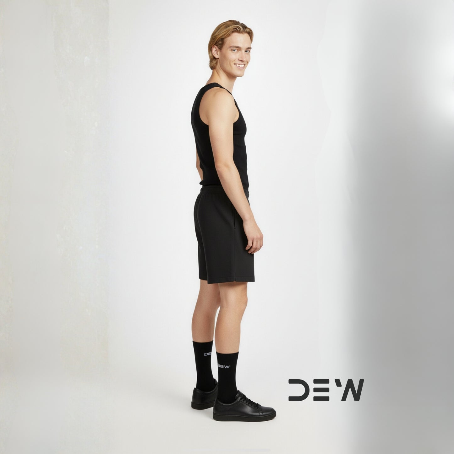 Person wearing a black sleeveless top and shorts with black Dew socks and shoes on a white background, featuring the brand 'DEW'.