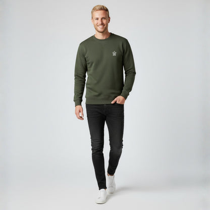 Man wearing a Dew olive green crewneck jumper with a logo, black jeans, and white shoes on a light gray background