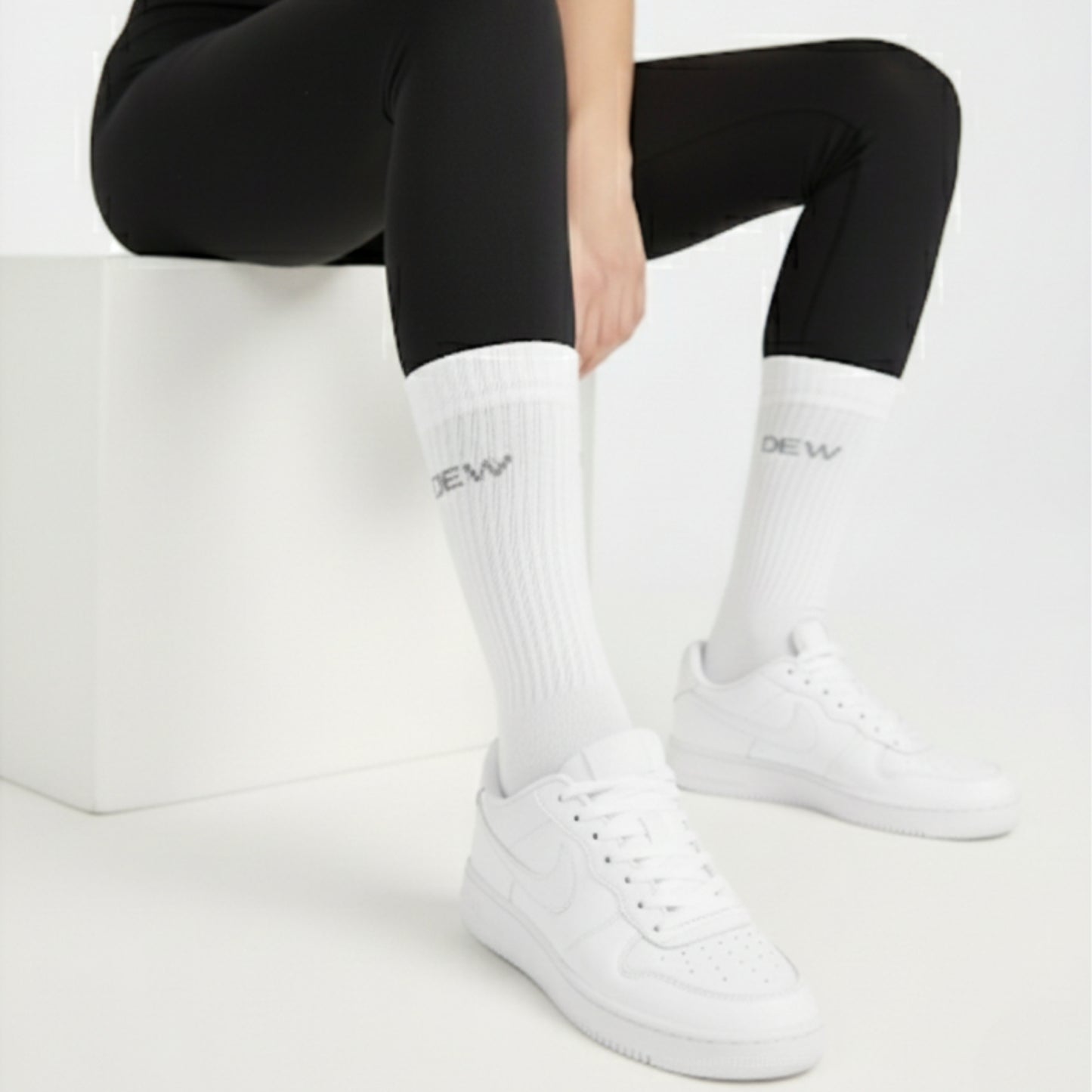Person wearing black leggings, white Dew socks with branding, and white sneakers on a light background