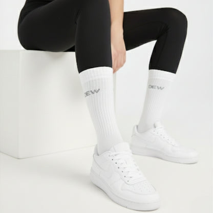 Person wearing black leggings, white Dew socks with branding, and white sneakers on a light background