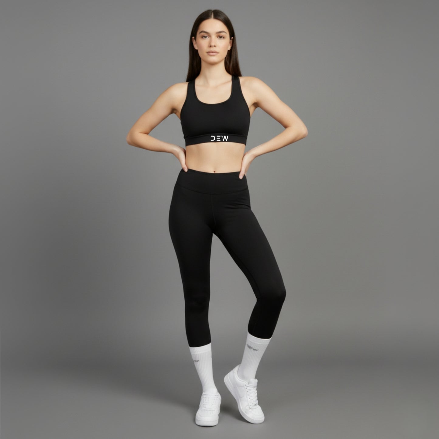 Woman wearing a black sports bra and leggings with white Dew socks and sneakers on a gray background