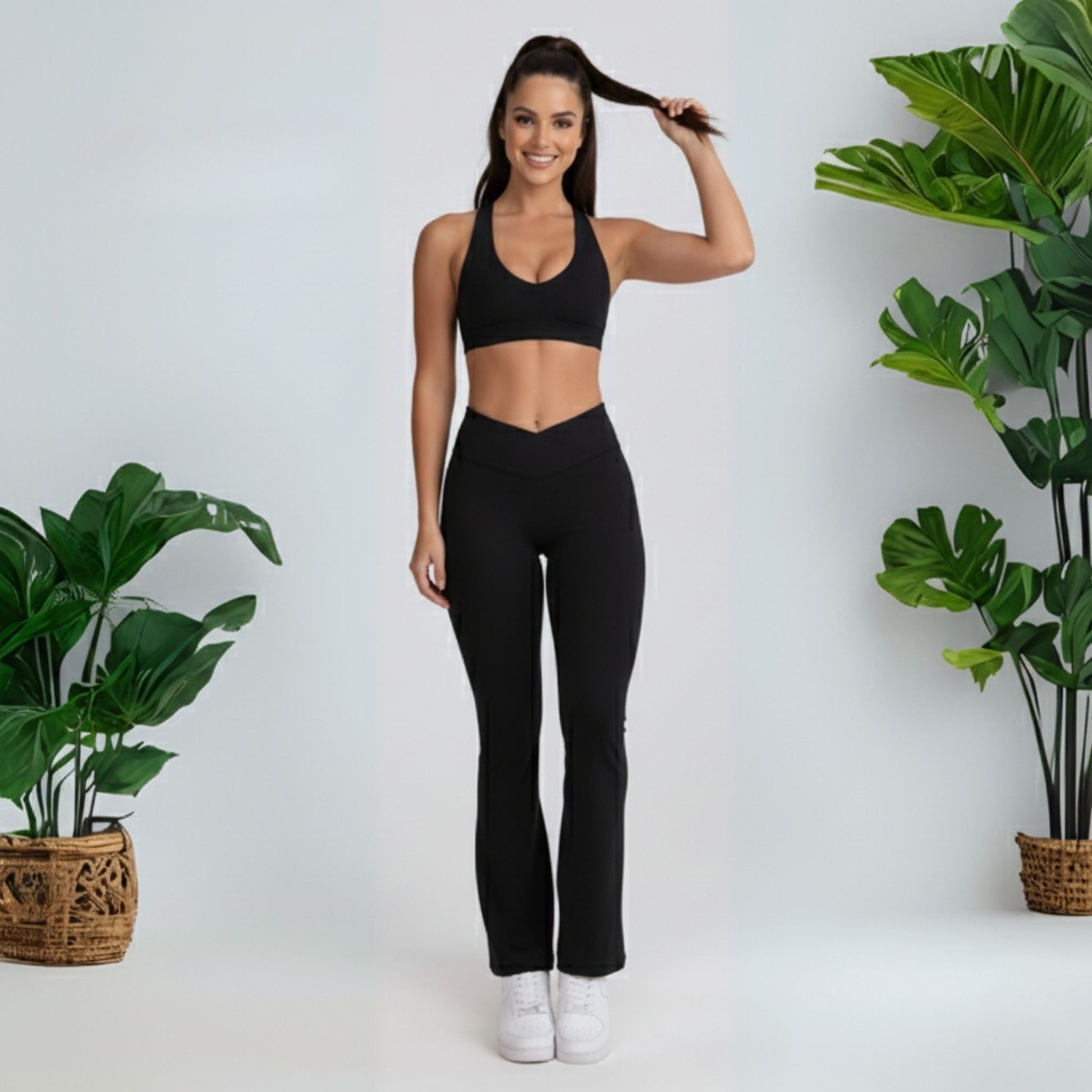 Woman in black  black dew flow fit flares  athletic wear posing against a white background with plants on either side.