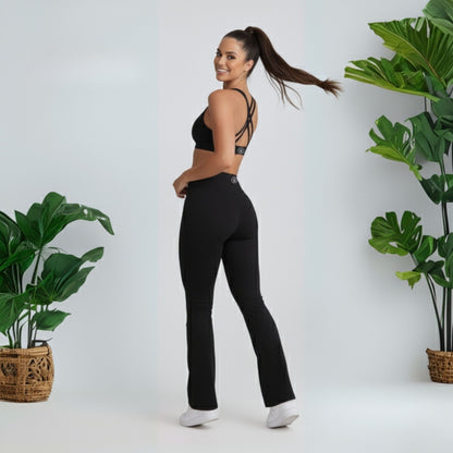 Woman wearing black  black dew flow fit flares  athletic pants and sports bra standing in a minimalistic setting with plants.