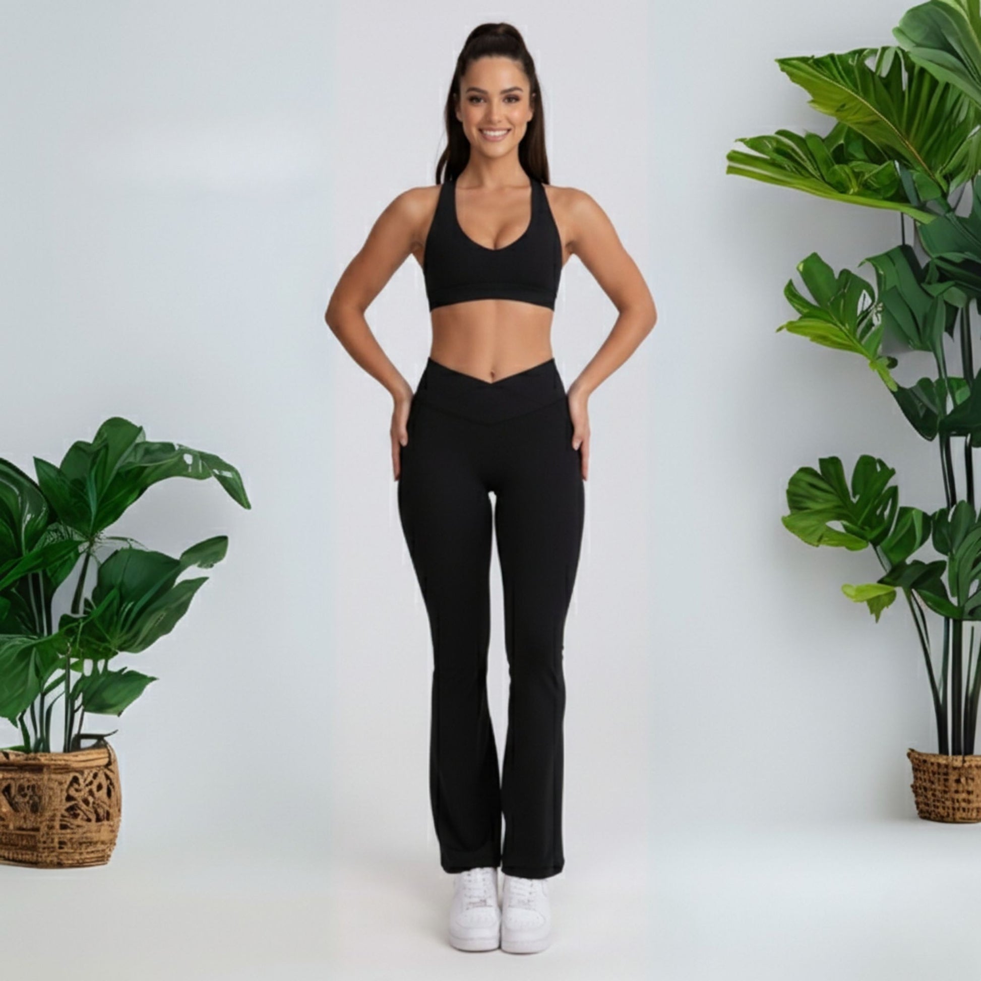 Woman in black dew flow fit flares athletic wear standing between two potted plants on a white background