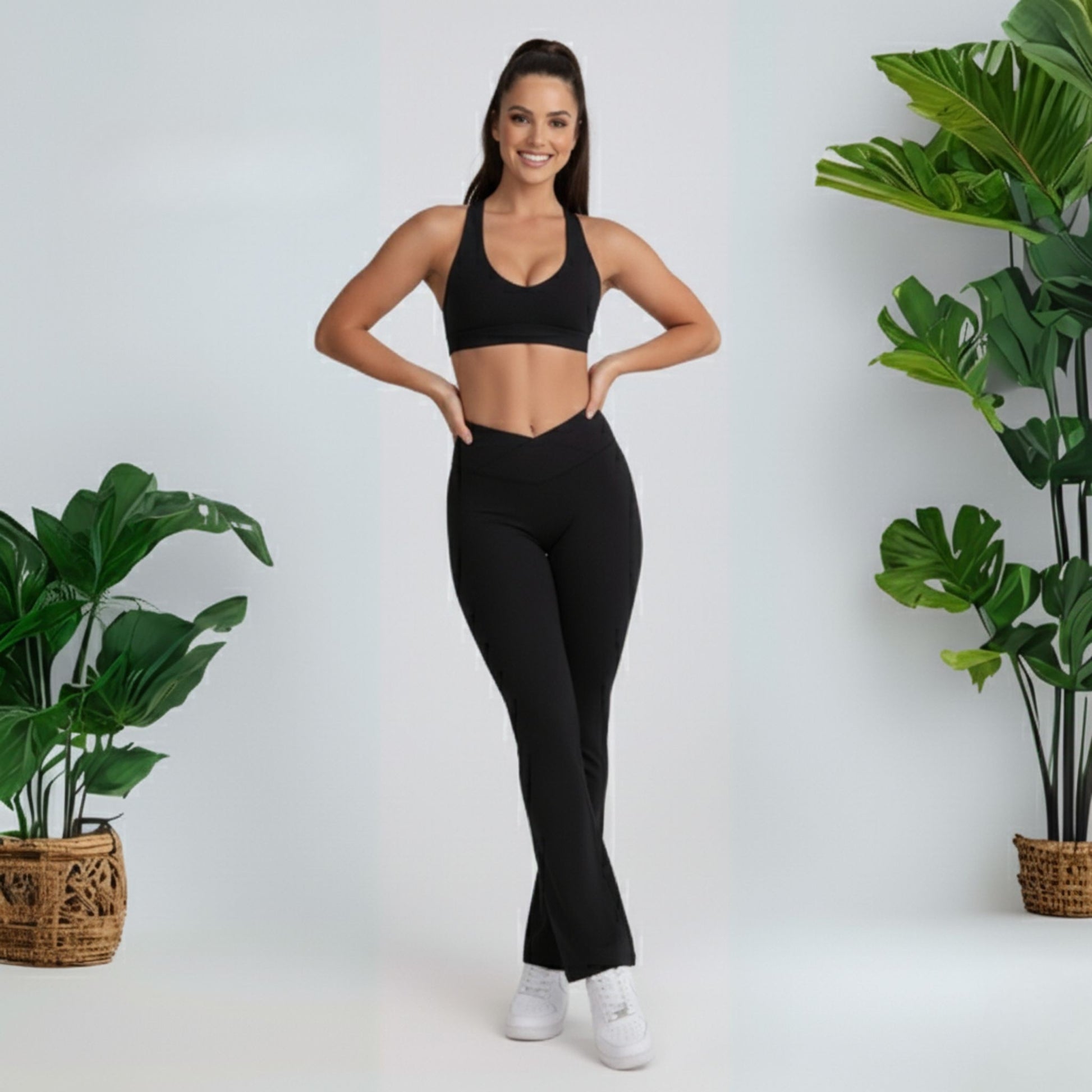 Woman in black  black dew flow fit flares  athletic wear standing between two potted plants on a white background