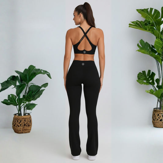 Woman wearing a  black dew flow fit flares sports bra and leggings set with plants on either side.
