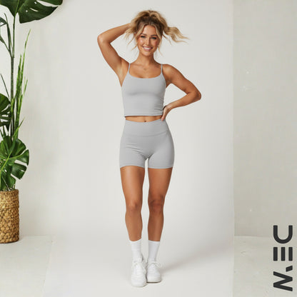 Woman in gray athletic wear posing in a studio setting with a plant and 'DEW' branding.